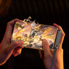 Hands hold a smartphone displaying a game, with a foldable cable charging the device.
