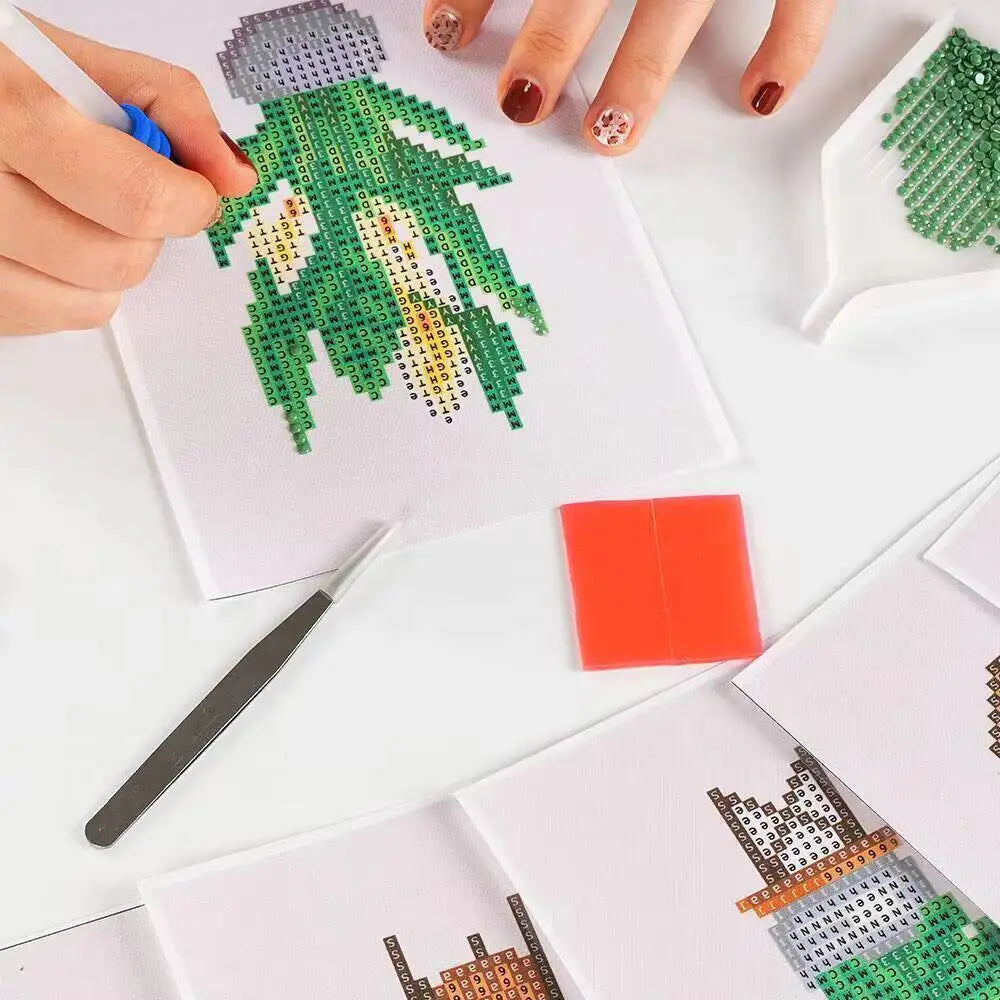 Hands are painting a green mountain landscape design on a white canvas with small colored beads.