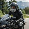 A man wearing a black leather jacket and helmet is riding a motorcycle on a road with mountains in the background.