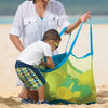 A woman is handing a large mesh beach and pool tote bag to a young boy on a sandy beach.