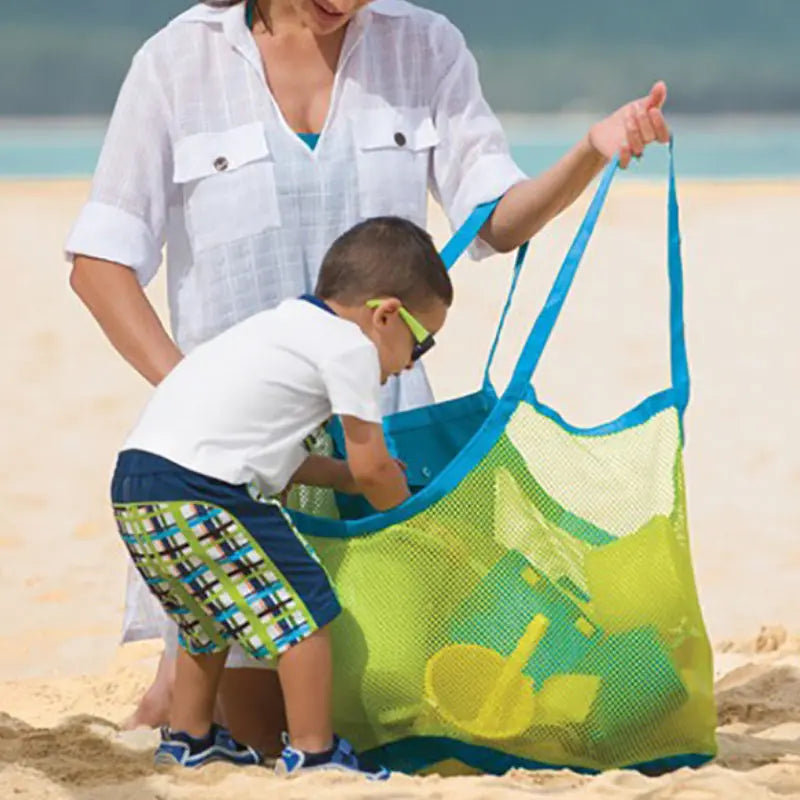 A woman is handing a large mesh beach and pool tote bag to a young boy on a sandy beach.