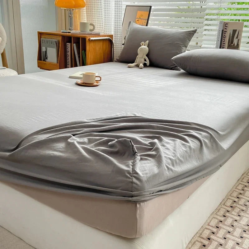 A fitted sheet in solid gray is draped over a bed, with pillows and wooden furniture visible.