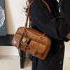 Eine Frau hält eine braune Handtasche mit mehreren Taschen und goldfarbenem Zierwerk vor weißem Hintergrund.