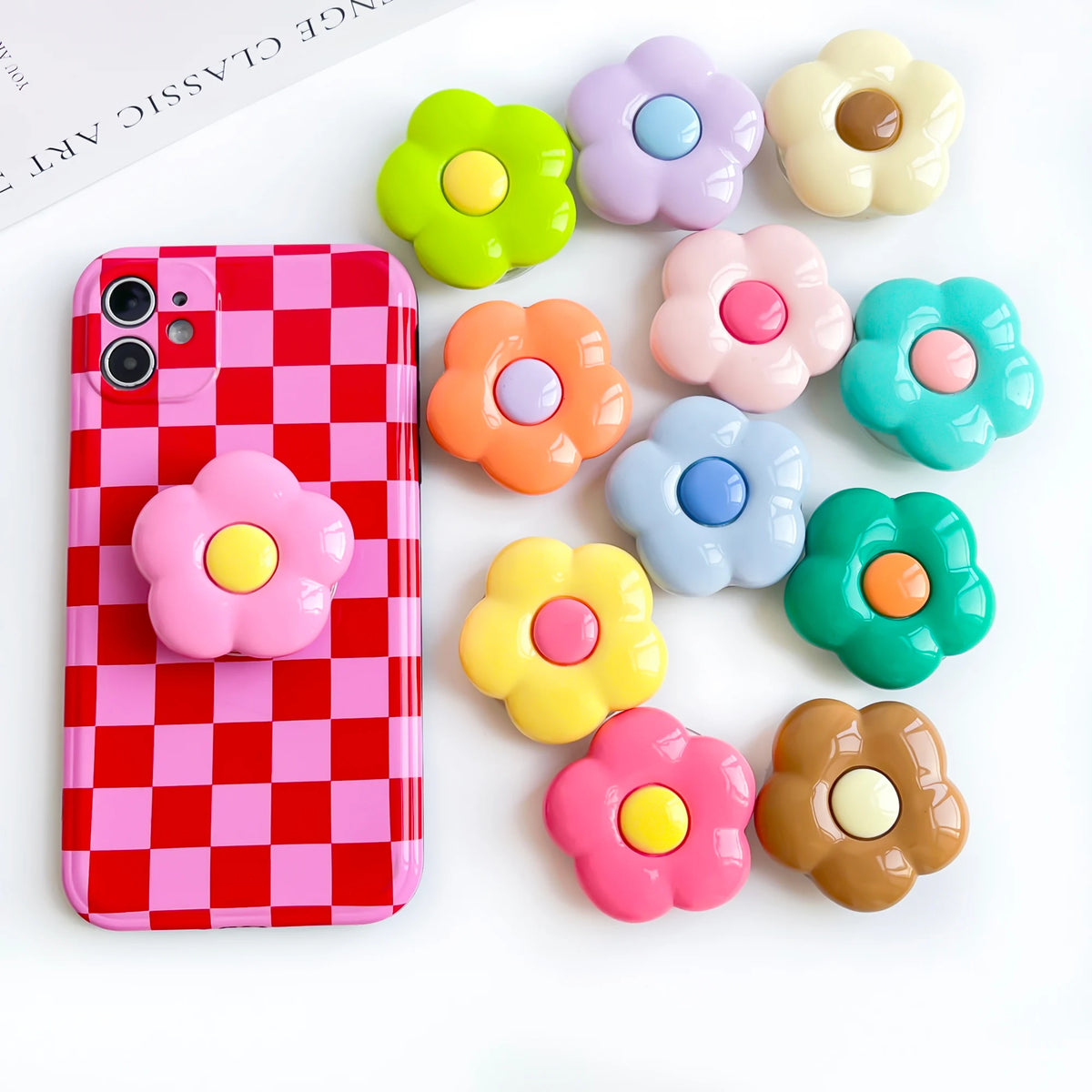 A pink checkered phone case is displayed next to several colorful, flower-shaped grips.