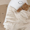A soft floral muslin baby blanket with a smiling face and a brown teddy bear toy.