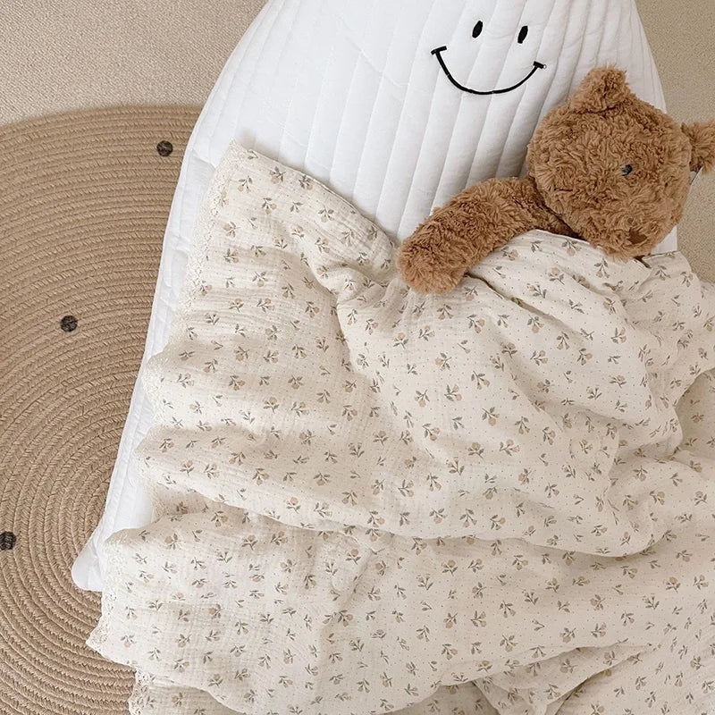 A soft floral muslin baby blanket with a smiling face and a brown teddy bear toy.