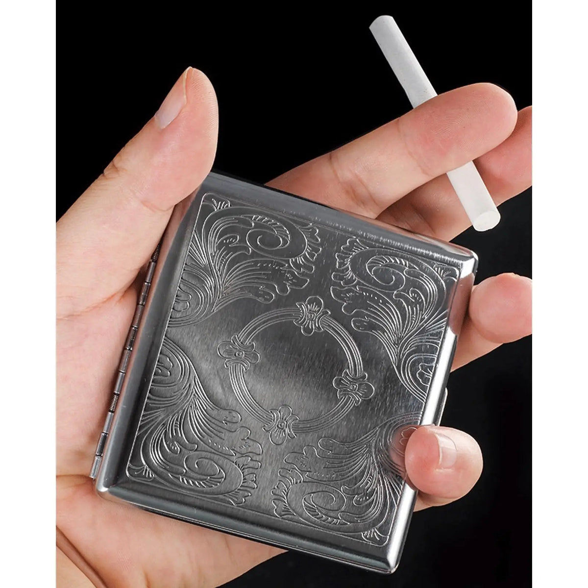A hand holds a classic stainless steel cigarette case with an embossed pattern and a white cigarette tip.