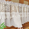 Beige sheer curtain with lace trim and tassel fringe hanging on a wooden shelf.