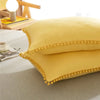 A yellow pillow with a pom-pom trim resting on a gray fabric surface.