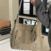 A hand holds a beige matte leather crossbody hobo bag with a silver zipper detail against a window background.