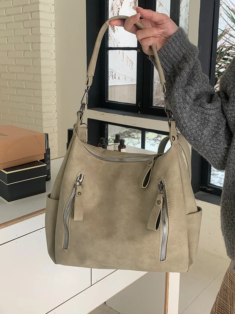 A hand holds a beige matte leather crossbody hobo bag with a silver zipper detail against a window background.
