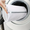 A hand is placing a white mesh shoe washing bag with a zipper closure into the drum of a washing machine.