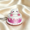A pink and white cartoon cake shaped like a cat is displayed on a satin cloth with a silver chain nearby.