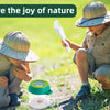 Two children explore nature, one holding a white handheld insect catcher clamp in a grassy outdoor setting.