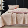 A floral quilted bedspread in soft beige and pink tones is draped over a dark headboard.