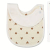 A light beige baby cotton gauze bib with a curved top and a scalloped edge, featuring small brown heart embroidery.