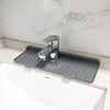 A silver silicone faucet splash mat with a spout is placed on a white countertop next to a sink.