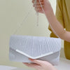 A hand holds a silver pleated glitter envelope clutch with a detachable chain strap.