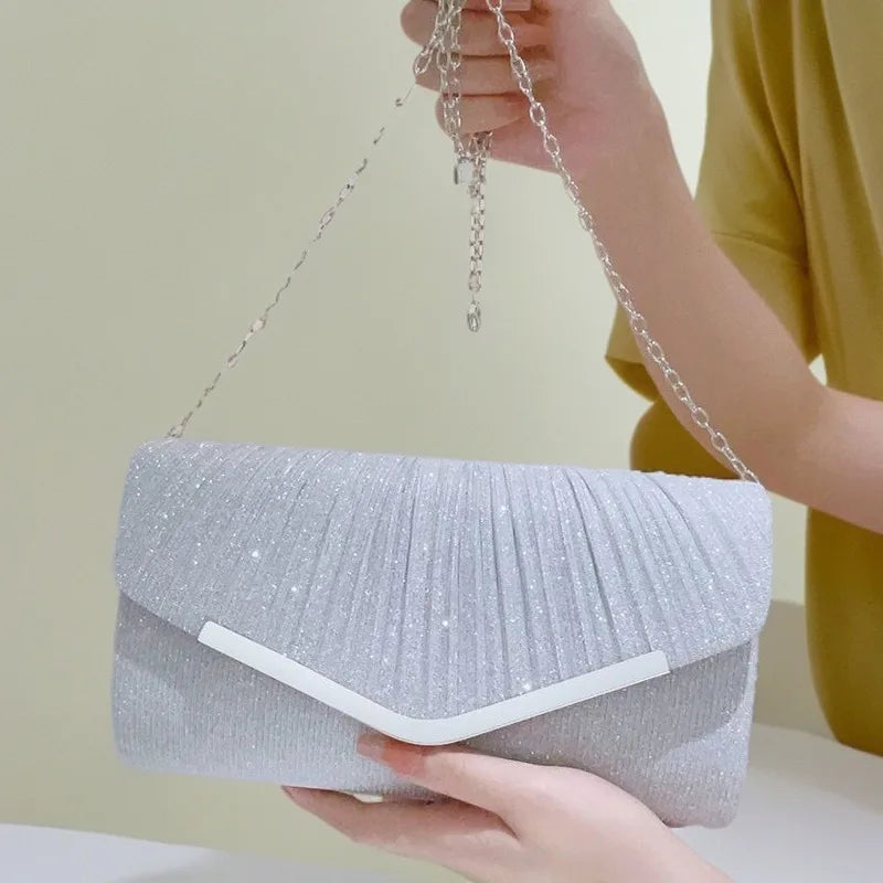 A hand holds a silver pleated glitter envelope clutch with a detachable chain strap.