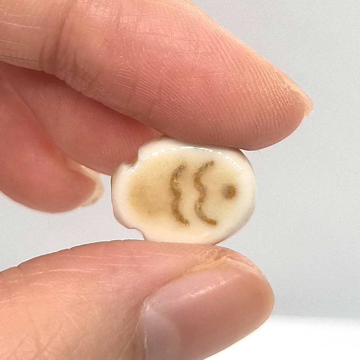 A small, light-colored ceramic fish bead with a stylized face design is held between fingers.