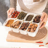 A clear plastic container filled with various dried spices and herbs, held by a person over a wooden board.