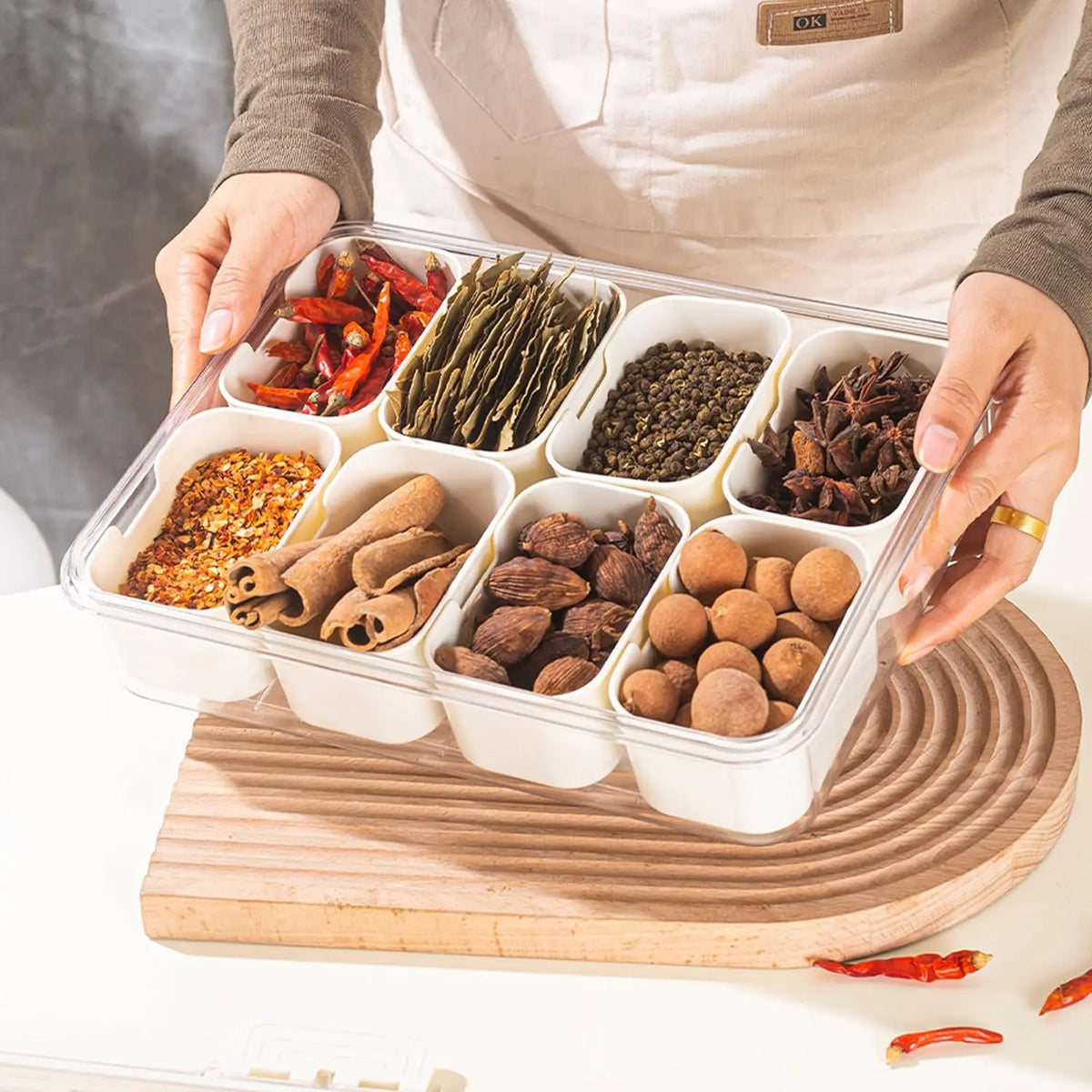 A clear plastic container filled with various dried spices and herbs, held by a person over a wooden board.