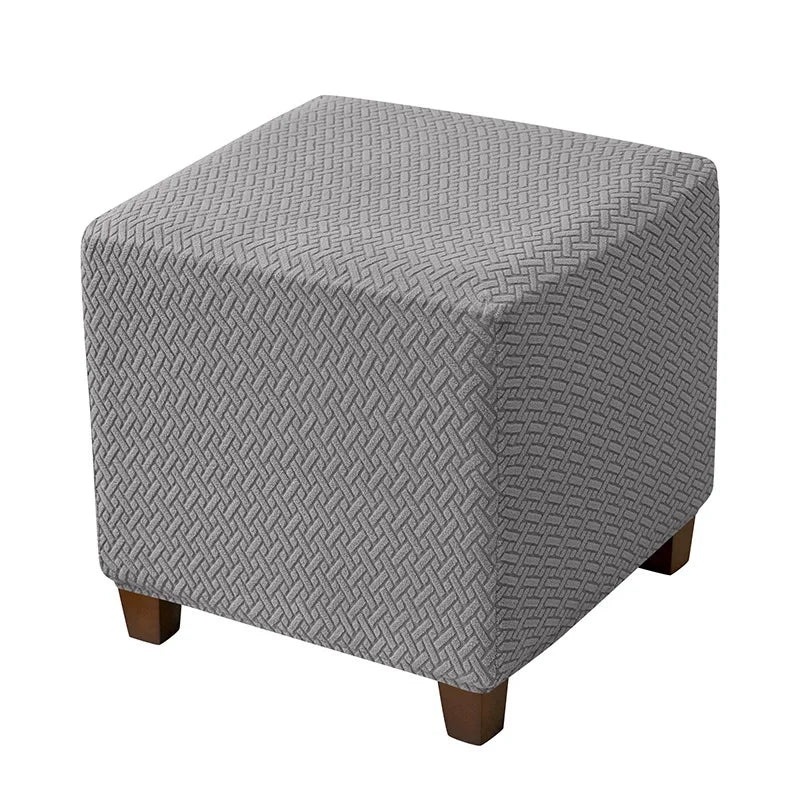 A square ottoman slipcover in a gray woven fabric with an elastic edge.