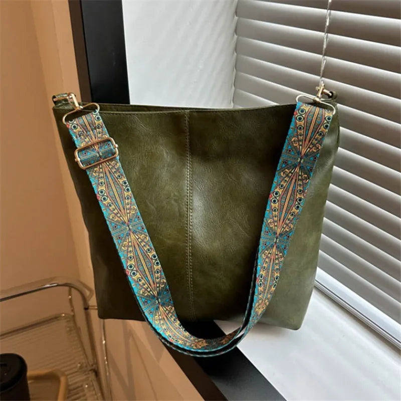 A dark olive green vintage bohemian shoulder bag with golden embroidery hanging in front of a window blind.