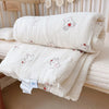 A white floral muslin baby blanket with a bear design is folded on a light-colored surface.