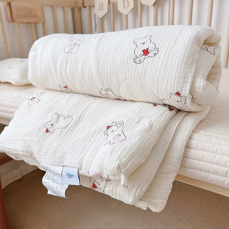 A white floral muslin baby blanket with a bear design is folded on a light-colored surface.