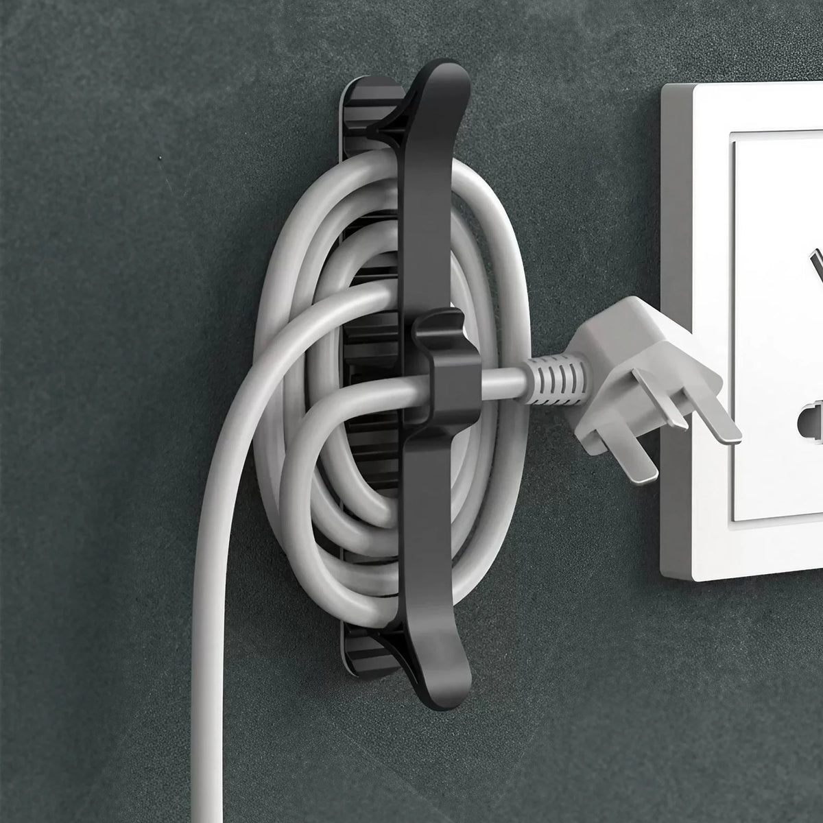 A white cable organizer with an adhesive mount is shown mounted on a dark gray wall next to a white electrical outlet.
