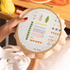 A hand holds a circular embroidery guide showing various hand embroidery stitches like fly, satin, and fern on a canvas.