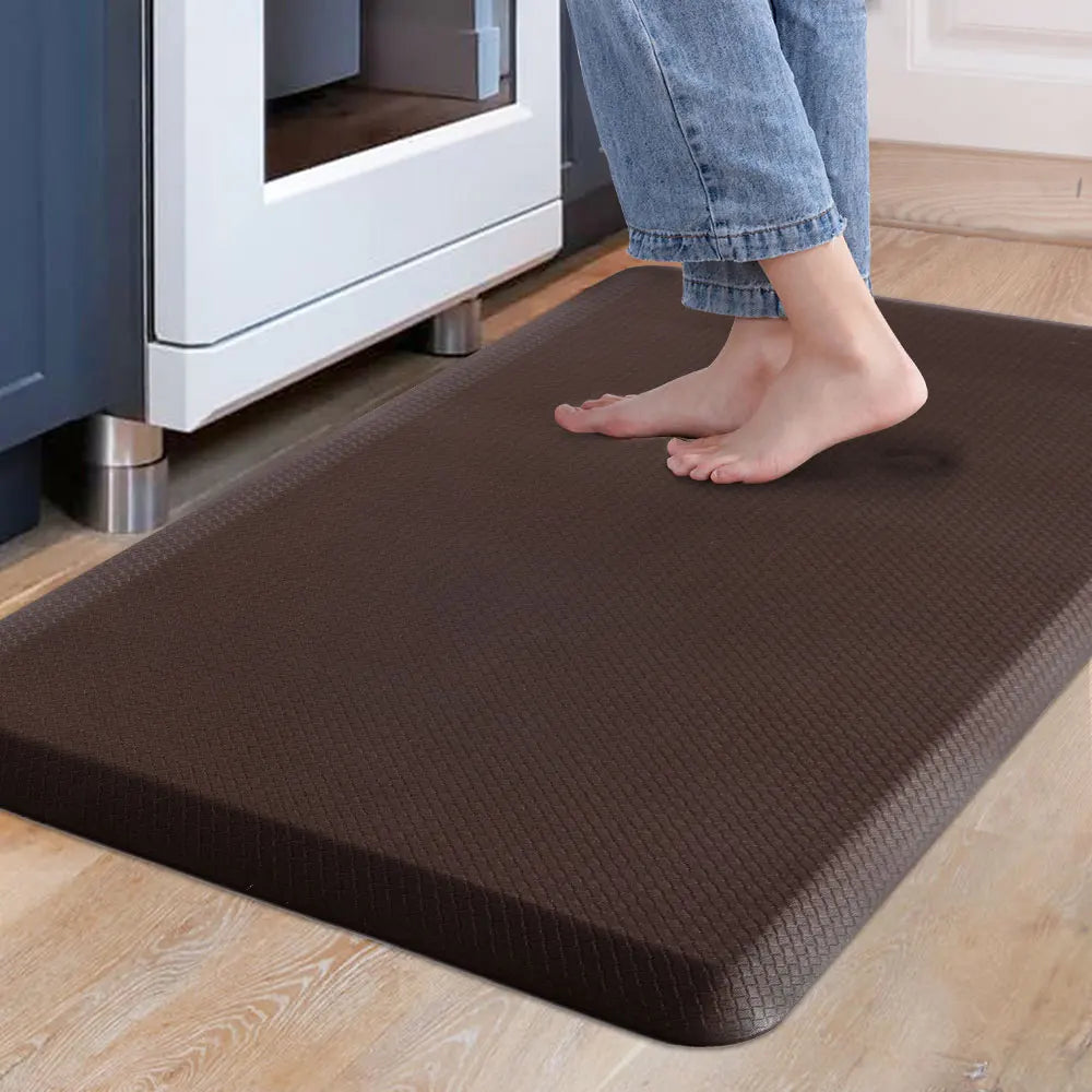 A person's feet are standing on a dark brown anti-fatigue mat placed on a light wooden floor near a white appliance.