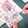 A hand is using precision craft tweezers to pick up a small paper cutout featuring a smiling girl and teddy bears.