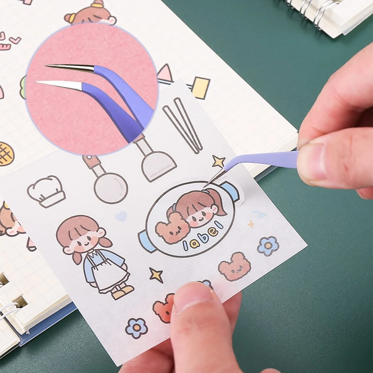 A hand is using precision craft tweezers to pick up a small paper cutout featuring a smiling girl and teddy bears.