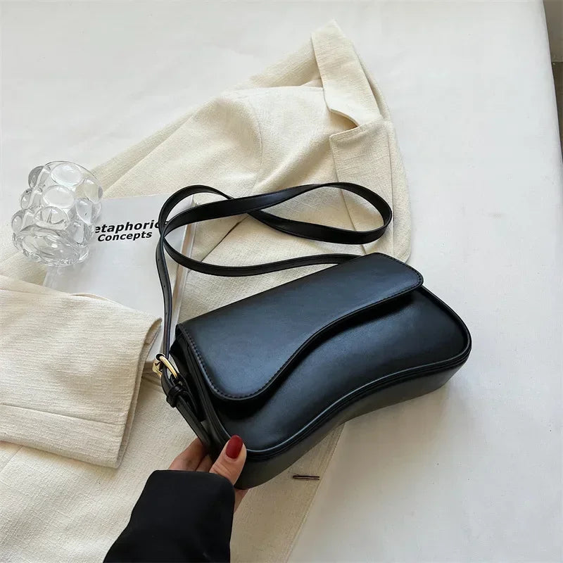 A black vintage leather crossbody bag with a flap closure is shown resting on a white surface next to a beige jacket.
