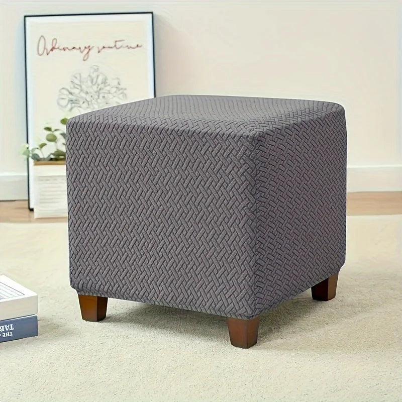 A gray stretch jacquard ottoman slipcover with an elastic edge sitting on a light carpet.