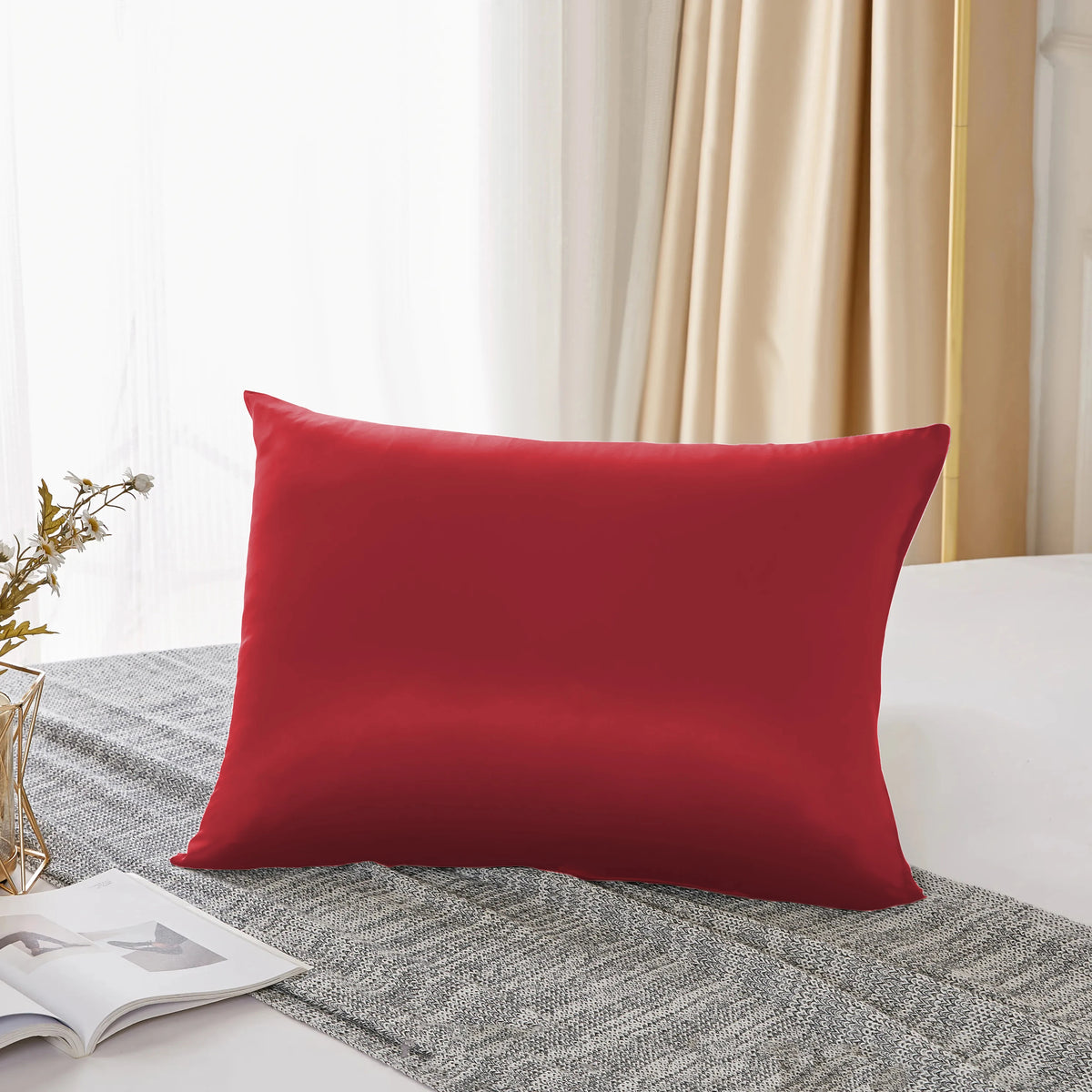 Une taie d'oreiller en soie rouge solide posée sur un tissu gris texturé près d'une fenêtre.