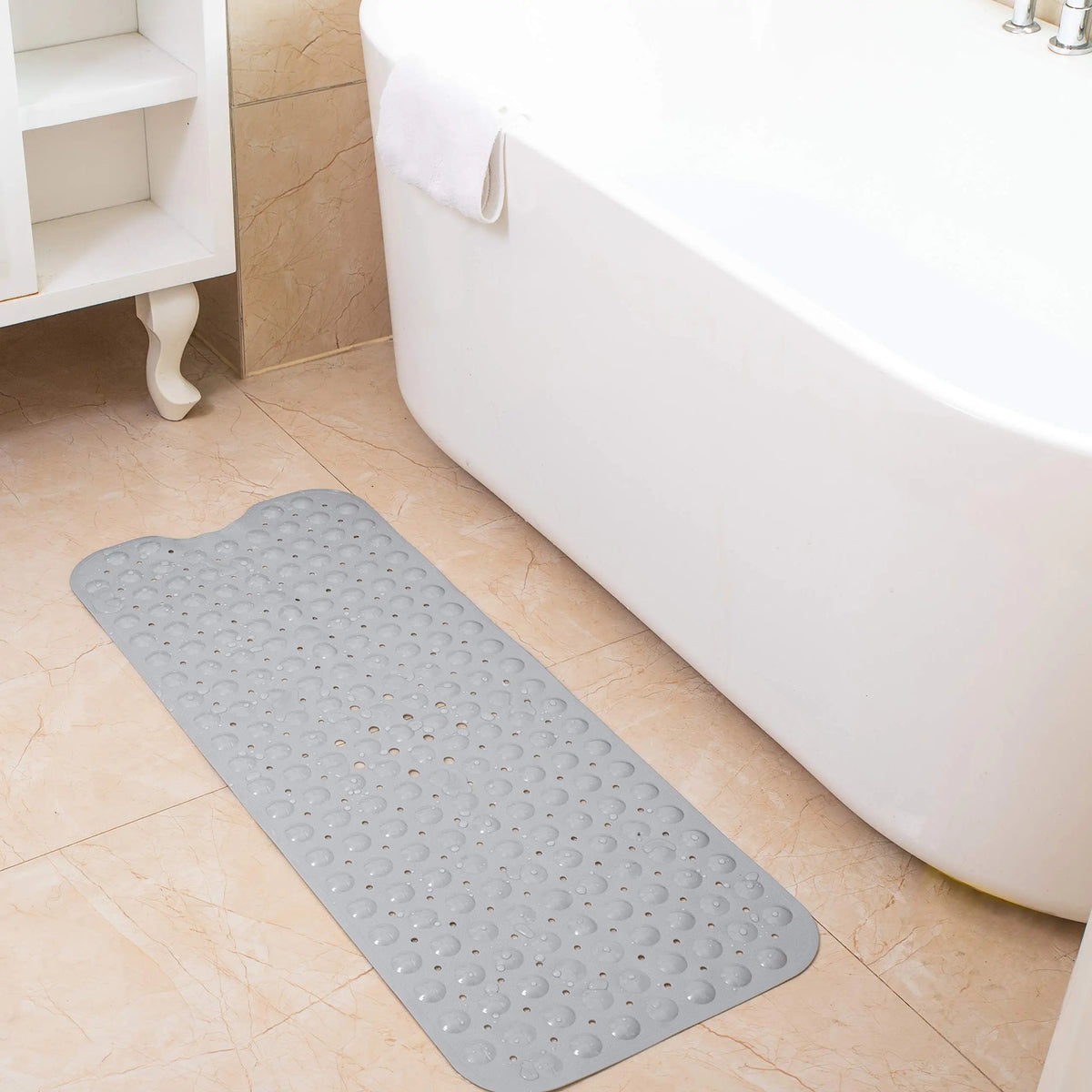A gray non-slip bathtub mat with suction cups and drain holes placed on a tiled floor next to a white bathtub.
