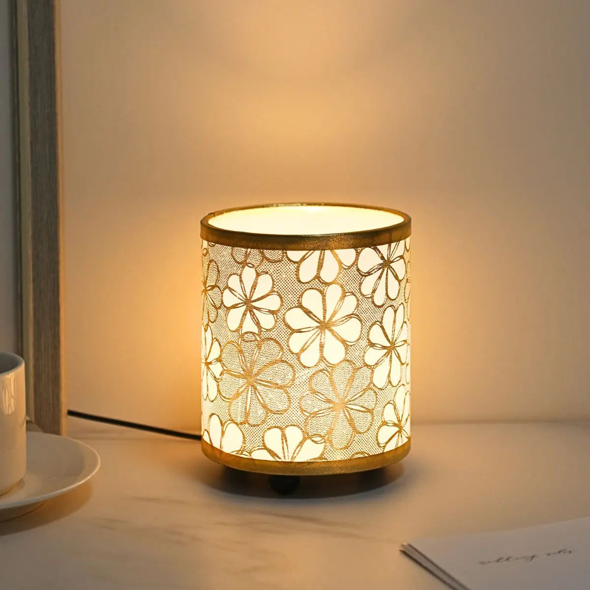 A gold-toned printed fabric USB desk lamp with a warm glow sitting on a white surface.