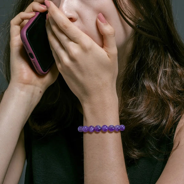 Eine Person hält ein pinkes Smartphone zum Mund, trägt ein lila Perlenarmband am Handgelenk.