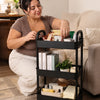 A woman is using a black 3-tier rolling utility cart to organize various items on a wooden floor.