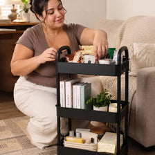 A woman is using a black 3-tier rolling utility cart to organize various items on a wooden floor.