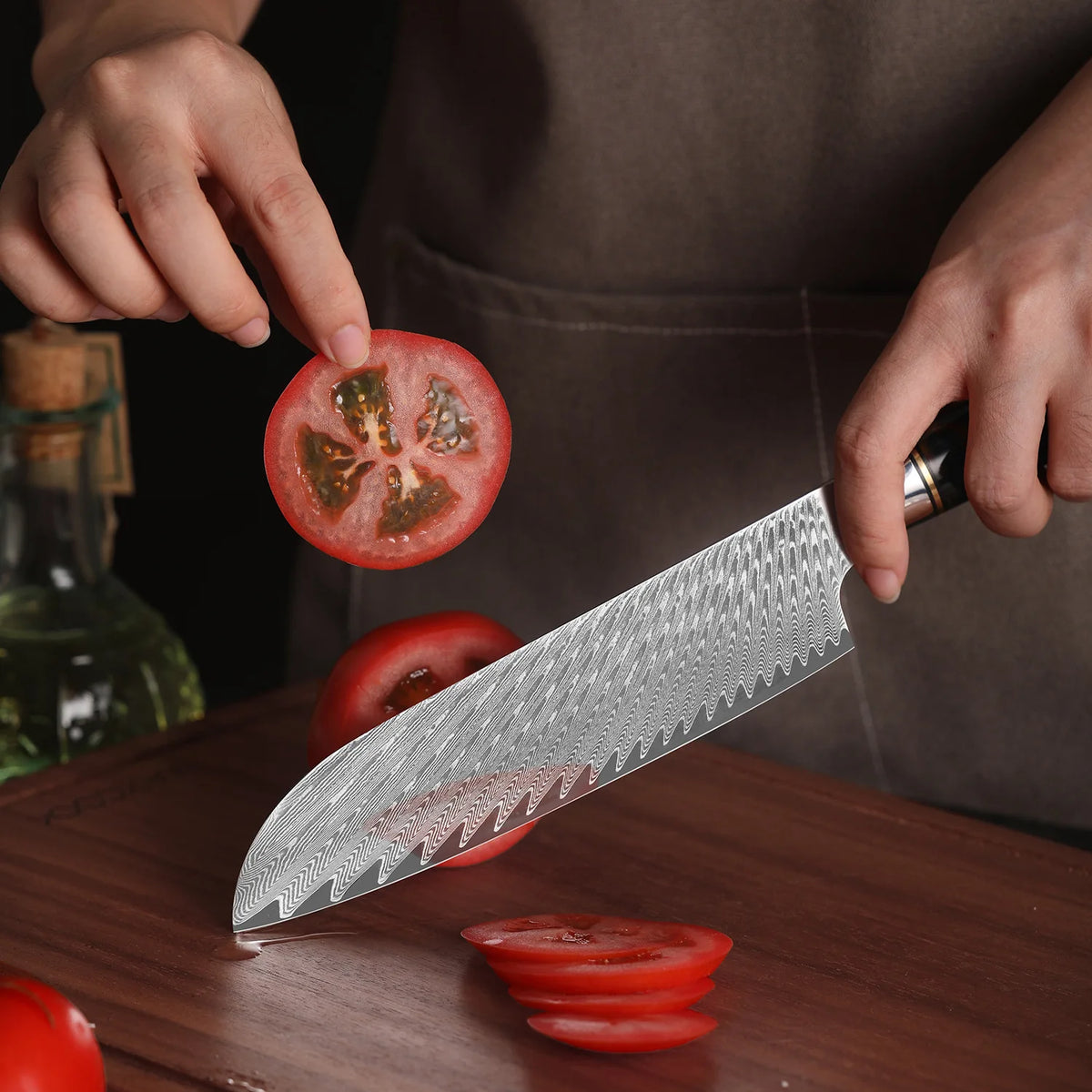 Hands slice a tomato on a wooden cutting board using a Damascus steel chef's knife.