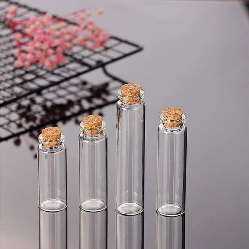 Five clear glass bottles with cork stoppers lined up on a surface, with pink glitter scattered in the background.