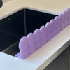 Lila Silikon-Spülschutzabfluss, platziert auf einer weißen Arbeitsplatte über einem schwarzen Waschbecken.