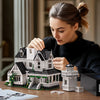 A woman is assembling a detailed Victorian house building kit with small wooden pieces on a table.
