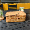 A woven rattan storage basket with a lid sits on a dark wooden surface next to a glass and yellow background.