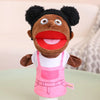 A soft plush hand puppet with dark curly hair, large eyes, and a wide red mouth, wearing a pink apron.
