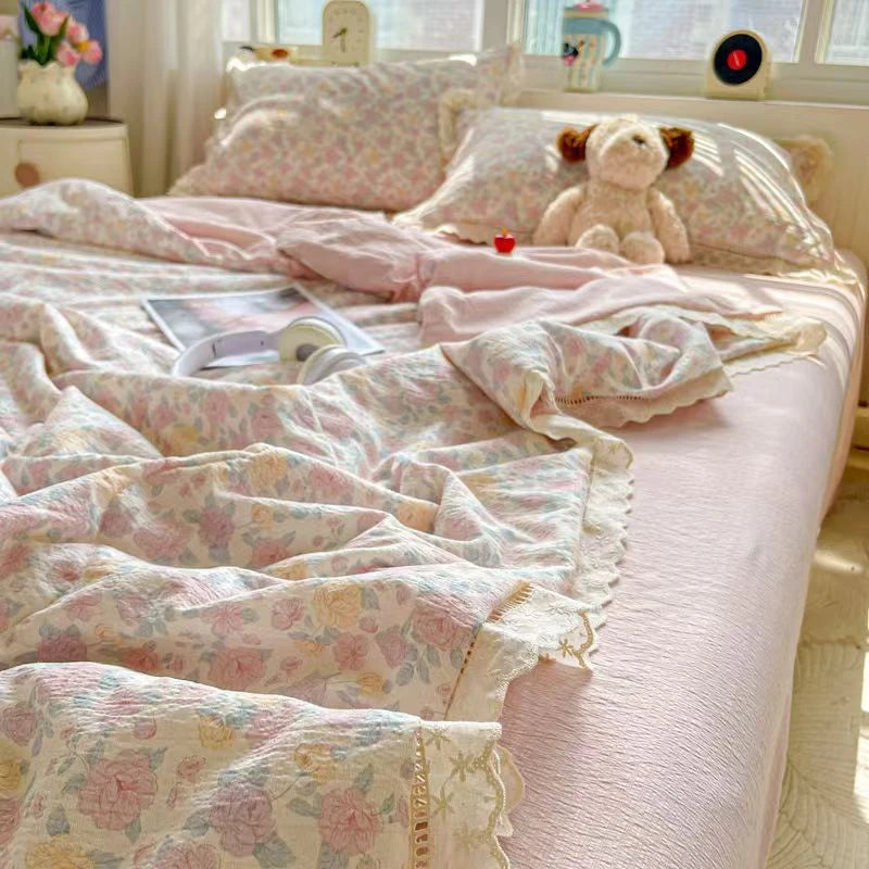 A pink floral washed cotton gauze summer quilt draped on a bed with pillows and a teddy bear.
